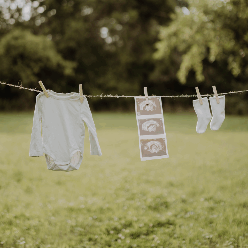 Leistungen nach der Geburt. Eine gespannte Wäscheleine über einer grünen Wiese mit einem aughängten Babybody, einem Abzug von 3 Ultraschallaufnahmen und einem paar weiße Babysocken.