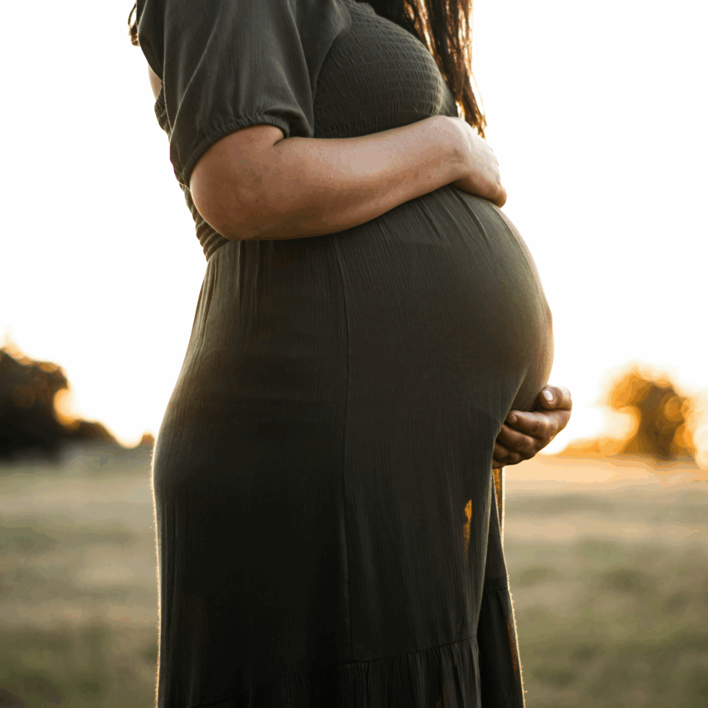 Leistungen zur Schwangerschaft und Geburtsvorbereitung. Frau hat Arme um schwangeren Bauch geschlungen, trägt dunkelgrünes Kleid und steht im Sonnenuntergang auf einer Wiese